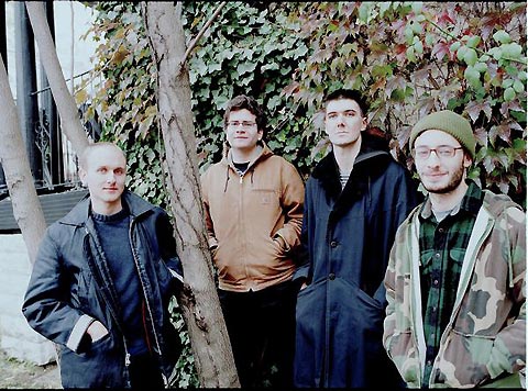 Four men in jackets stand next to a tree.