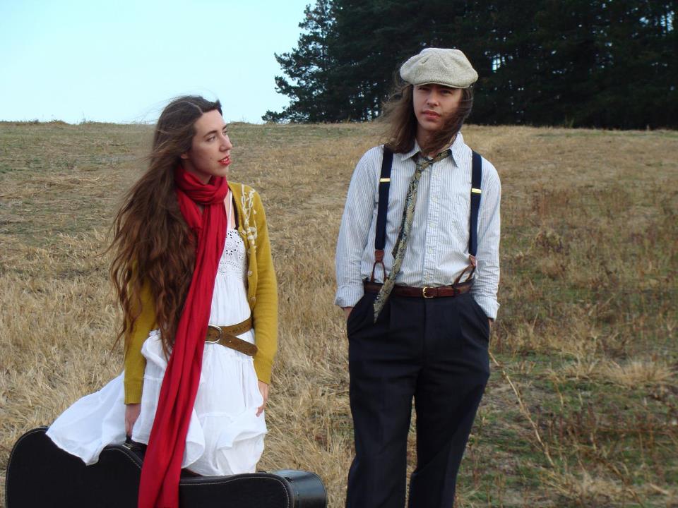 A man and woman standing in a field, she is holding a guitar case.