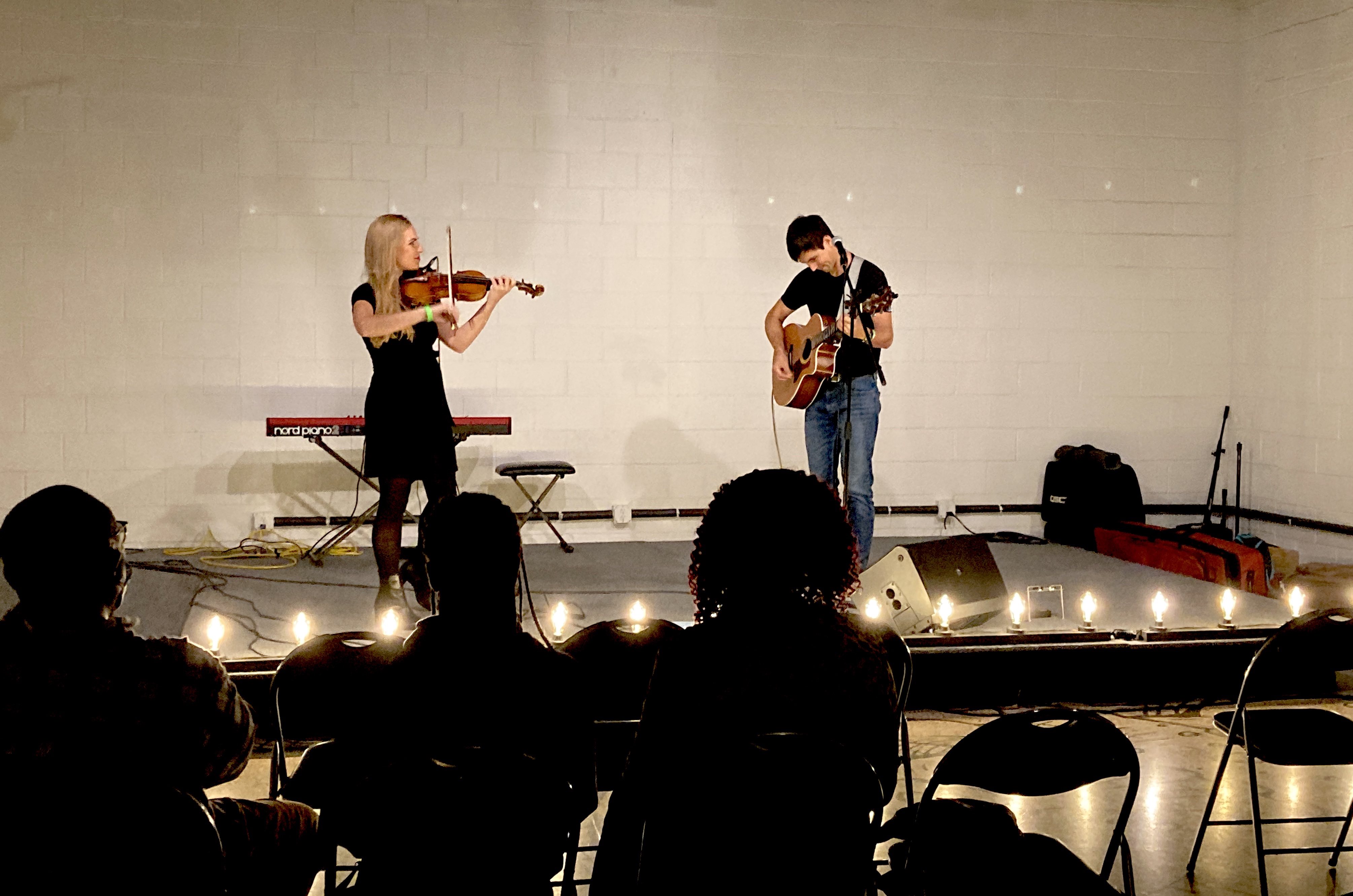 Two musicians play violin and acoustic guitar on a stage with minimal lighting, lined with Edison bulbs