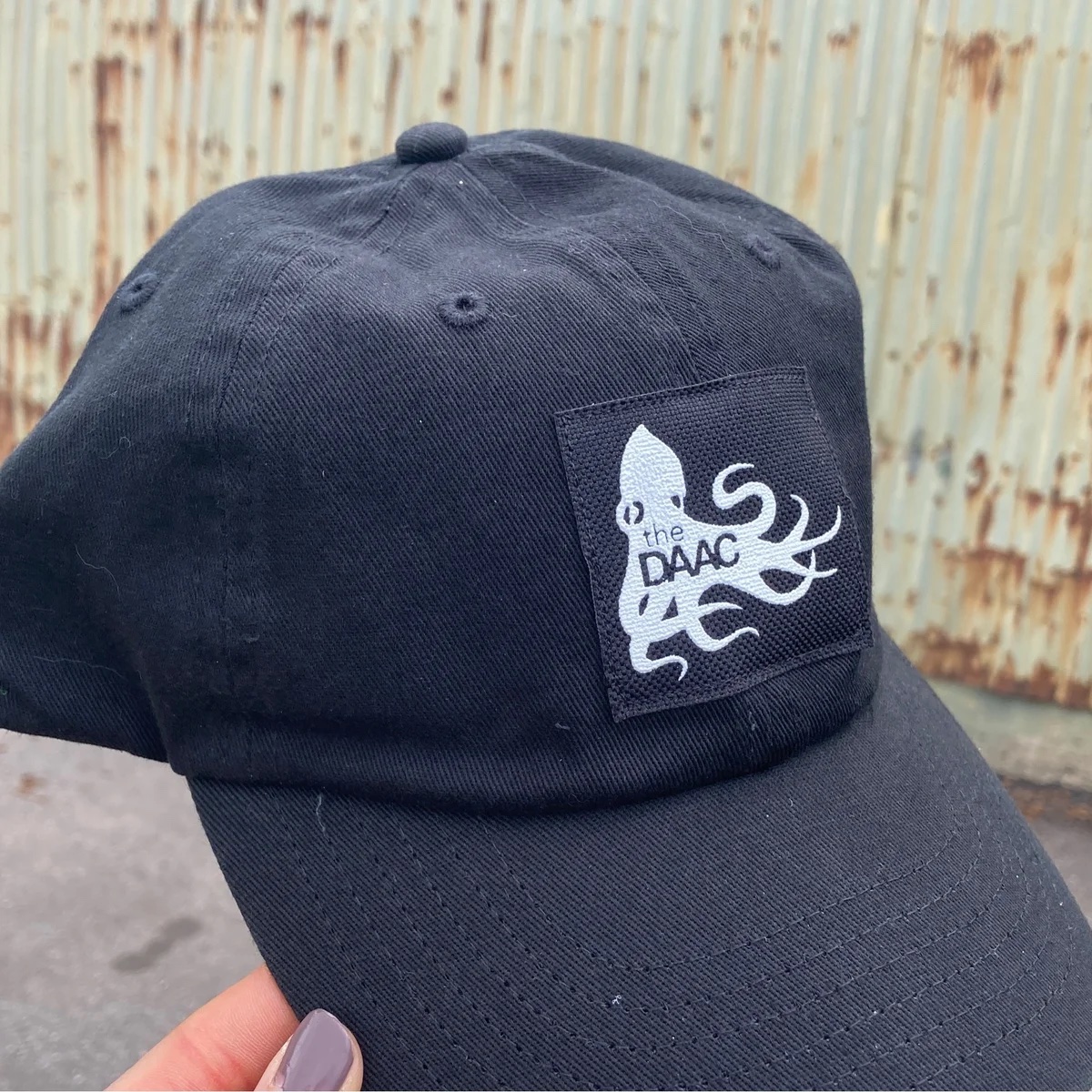 Black baseball cap with a printed black patch with white DAAC logo on the front