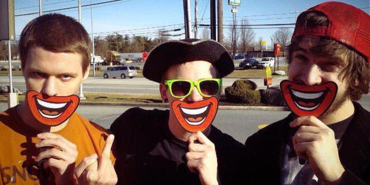 Three men standing on the street holding smiley face signs in front of their mouths