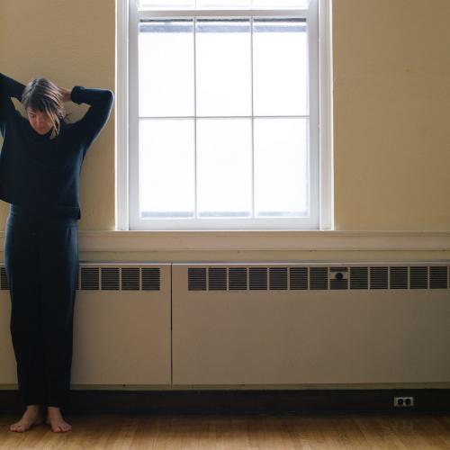 A person with straight, sandy brown hair stands next to a beige wall and window