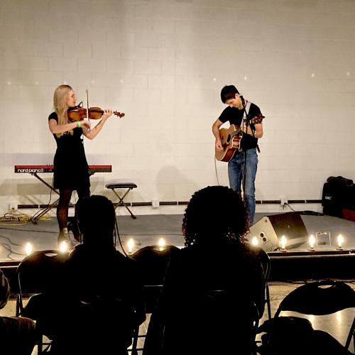 Two musicians play violin and acoustic guitar on a stage with minimal lighting, lined with Edison bulbs